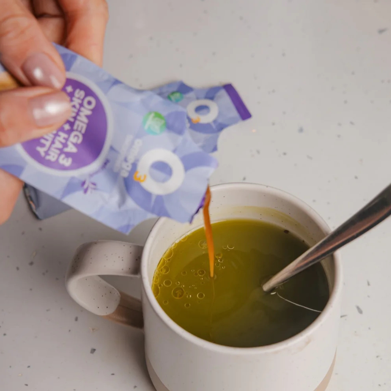 Woman pouring liquid O3Omega Skin, Hair & Immunity supplement from a single-serve sachet into a matcha green tea mug.